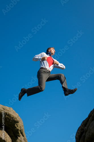 Superhero businessman taking a daring leap between two boulders outdoors against bright blue sky