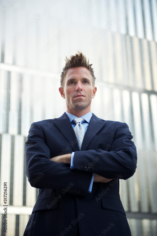 Serious young businessman standing outdoors with arms crossed in front of a modern urban skyscraper background Serious young businessman standing outdoors with arms crossed in front of a modern urban skyscraper background