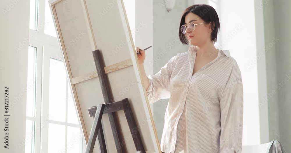 Portrait of paintress is painting picture on easel standing near the window, bottom view. Workflow process of creative drawing work of young woman artist in glasses on workplace in modern art studio.