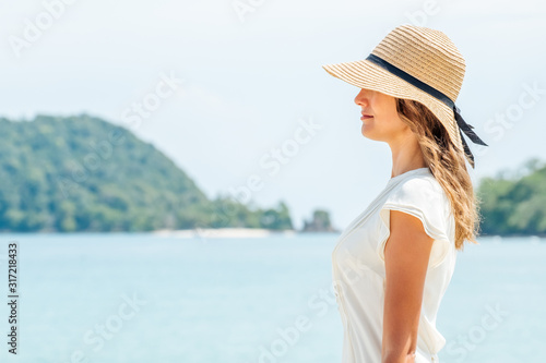 Beautiful tanned woman in white dress covering face with a straw hat © Iurii