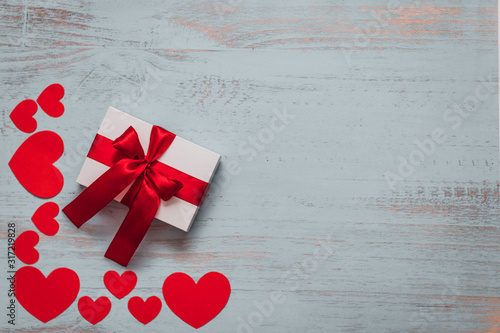 Paper hearts and a white present with red ribbon on a light painted wooden background. Top side angle view, flat lay. Valentines day concept. Copyspace.