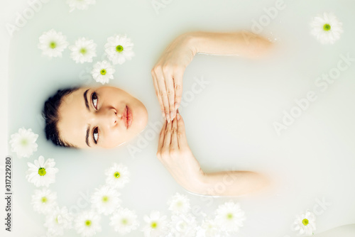 Happy young woman is lying in a milk bath with flowers, top view