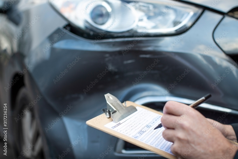 Close Up Of Auto Workshop Mechanic Inspecting Damage To Car And Filling In Repair Estimate Close Up Of Auto Workshop Mechanic Inspecting Damage To Car And Filling In Repair Estimate