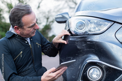 Auto Workshop Mechanic Inspecting Damage To Car And Filling In Repair Using Digital Tablet