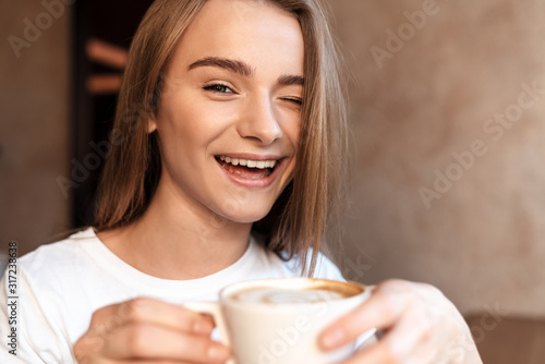 Photo of joyful young woman drinking coffee and winking