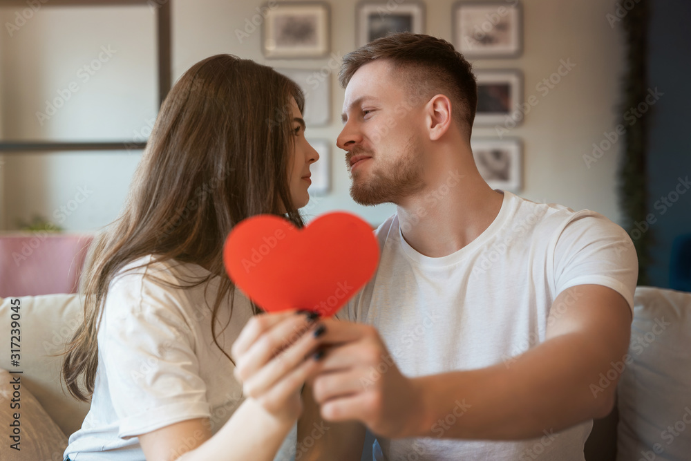 young romantic couple kissing on the sofa, both holding big red paper heard on saint valentine's day, happy unniversary young romantic couple kissing on the sofa, both holding big red paper heard on saint valentine's day, happy unniversary