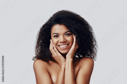 Cute young African woman smiling and looking at camera while standing against grey background
