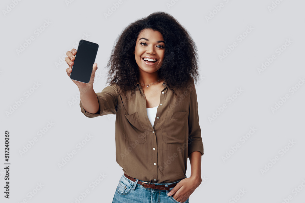 Beautiful young African woman pointing copy space at smart phone and smiling while standing against grey background Beautiful young African woman pointing copy space at smart phone and smiling while standing against grey background