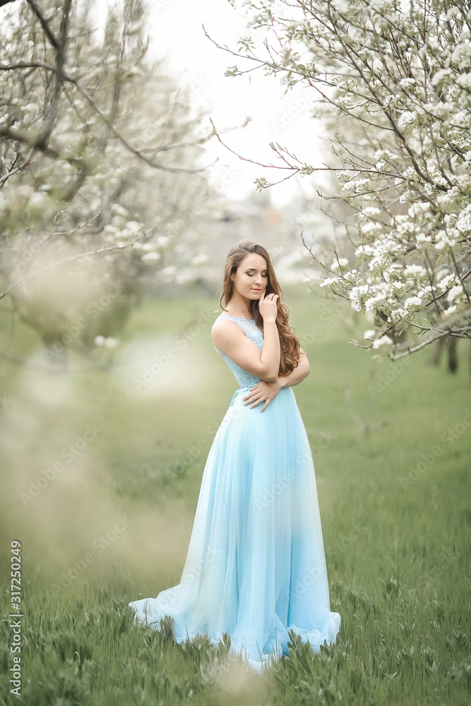 A young stylish model girl in a beautiful fashionable long green dress stands in a summer park near lush flower bushes and looks back.