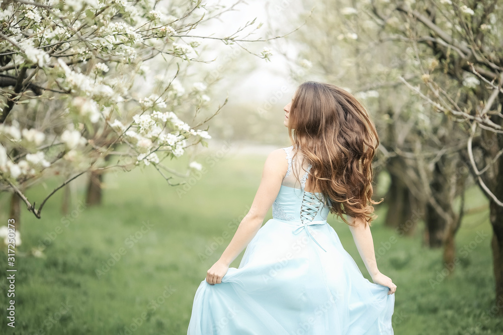A young stylish model girl in a beautiful fashionable long green dress stands in a summer park near lush flower bushes and looks back. A young stylish model girl in a beautiful fashionable long green dress stands in a summer park near lush flower bushes and looks back.