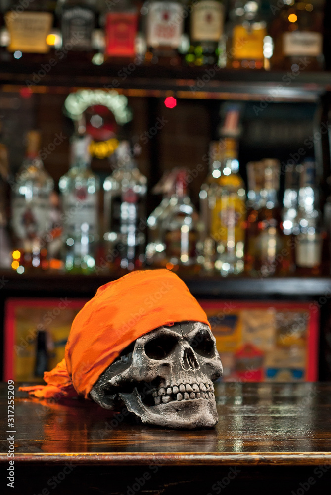 Human skull on a bar counter. Red skull in orange bandana on the background of bottles with alcohol. Biker Pub.