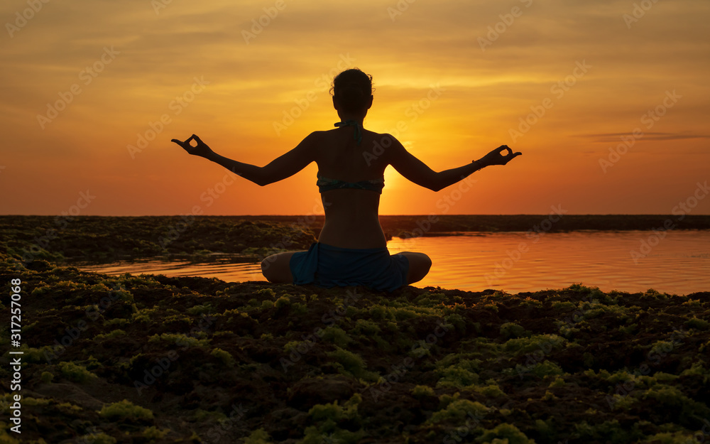 Yoga pose. Woman sitting on the beach, practicing yoga. Young woman raising arms with gyan mudra during sunset golden hour. View from back. Melasti beach, Bali. Yoga pose. Woman sitting on the beach, practicing yoga. Young woman raising arms with gyan mudra during sunset golden hour. View from back. Melasti beach, Bali.