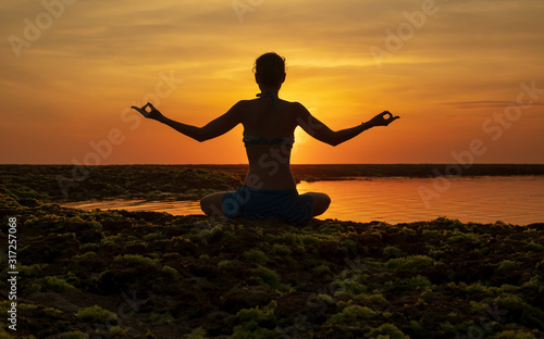 Yoga pose. Woman sitting on the beach, practicing yoga. Young woman raising arms with gyan mudra during sunset golden hour. View from back. Melasti beach, Bali.