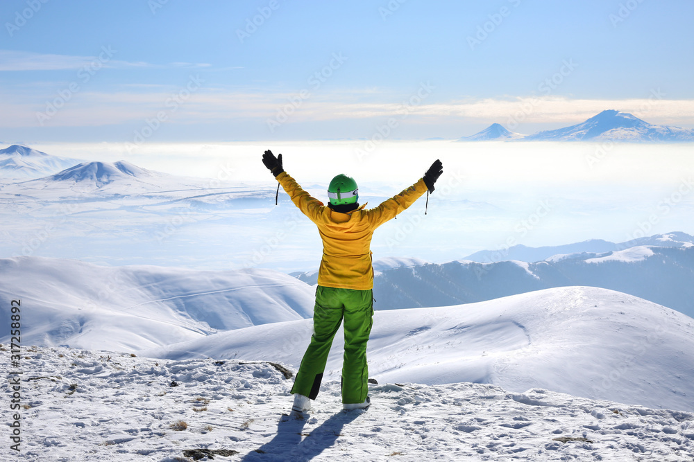 Happy woman in yellow ski jacket standing with hands up on the pick of mountain Tegenis. View of Ararat mountain.