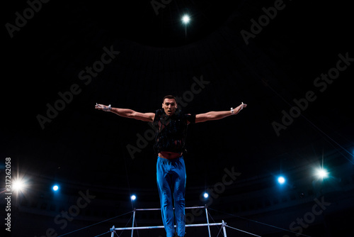athletic gymnast with outstretched hands performing near horizontal bars in arena of circus