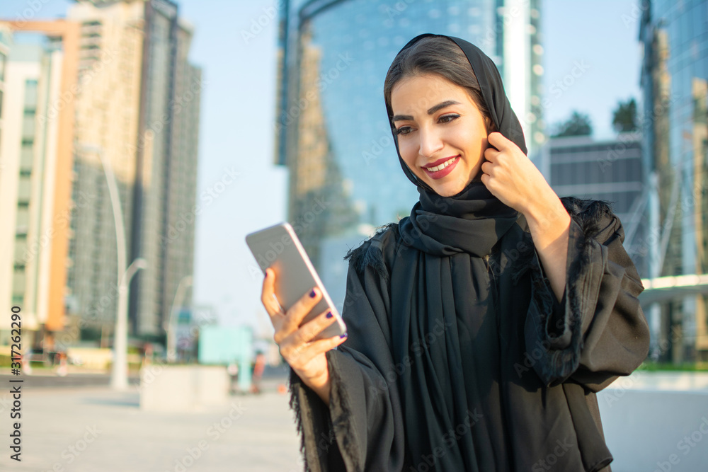 Beautiful smiling young Arabic woman in traditional abaya clothes using smartphone on the street. Beautiful smiling young Arabic woman in traditional abaya clothes using smartphone on the street.