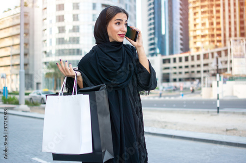 Arab woman in traditional wear holding shopping bags in hands and talking on mobile phone while standing on the street in front of the modern skyscrapers