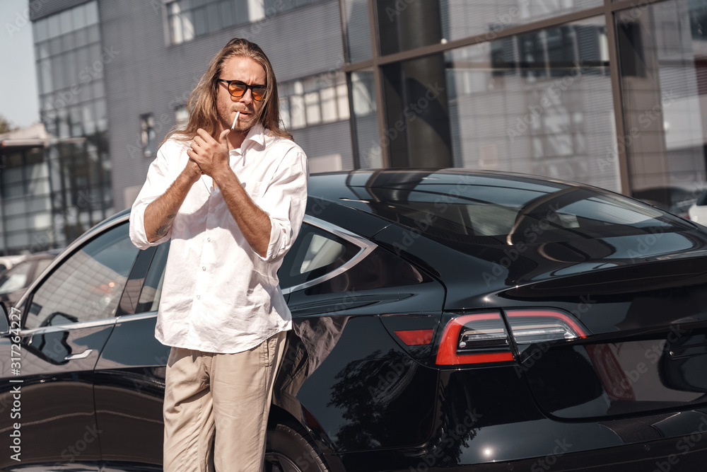 Fototapeta premium Mode of Transport. Man with long hair in sunglasses standing near electric car lighting cigarette looking aside cool