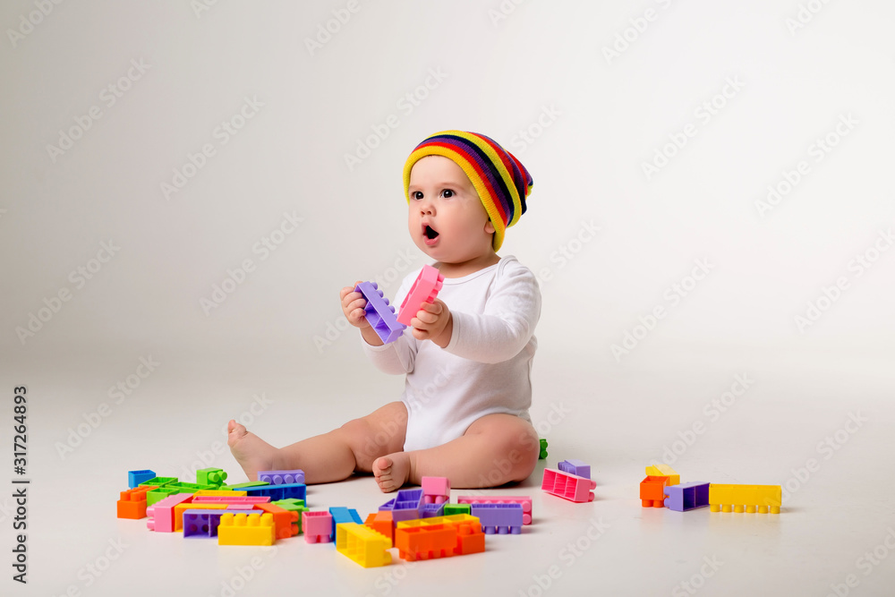 baby boy in white bodysuit and multicolored hat playing with a multicolored plastic constructor on a white background isolate, child development concept, space for text baby boy in white bodysuit and multicolored hat playing with a multicolored plastic constructor on a white background isolate, child development concept, space for text