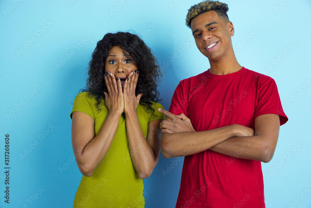 She's scared, he's laughting. Young emotional african-american beautiful man and woman in colorful clothes on blue background. Concept of human emotions, facial expession, relations, ad, friendship. She's scared, he's laughting. Young emotional african-american beautiful man and woman in colorful clothes on blue background. Concept of human emotions, facial expession, relations, ad, friendship.