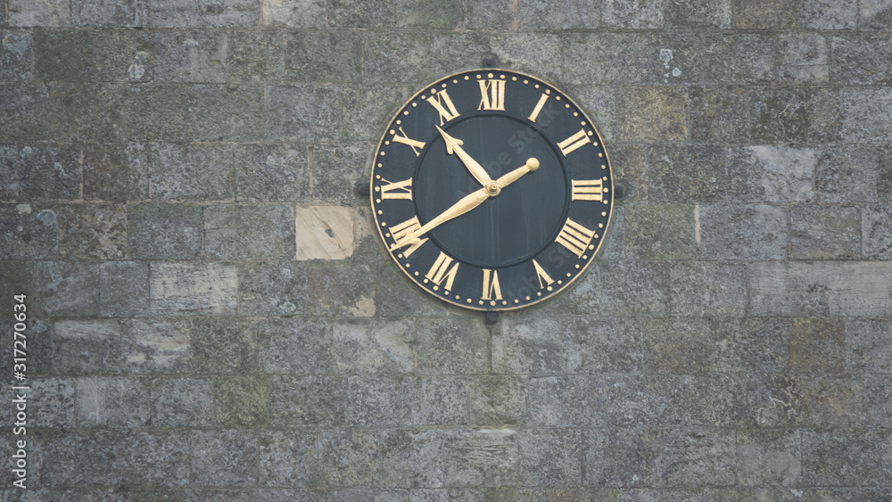 Black clock with gold hands and numerals on stone wall Black clock with gold hands and numerals on stone wall