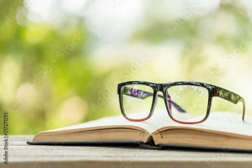 Book and eye glasses on wooden table with abstract green nature blur background. Reading and education concept