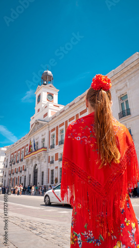 A young caucasian woman chulapa with red flower, traditional dress, and Spanish scarf at Puerta del Sol during San Isidro, the spring festival in May in the downtown of Madrid, the capital of Spain photo