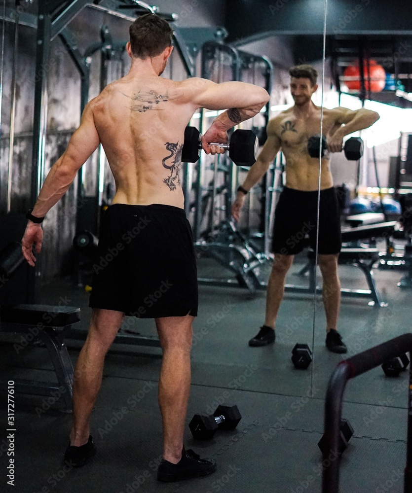 Fototapeta premium Caucasian Shirtless Tattooed Man Exercising Traps Using Dumbbells