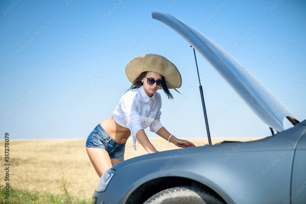 Naklejka premium Pretty young woman looking at the engine of broken car. Road trip problems