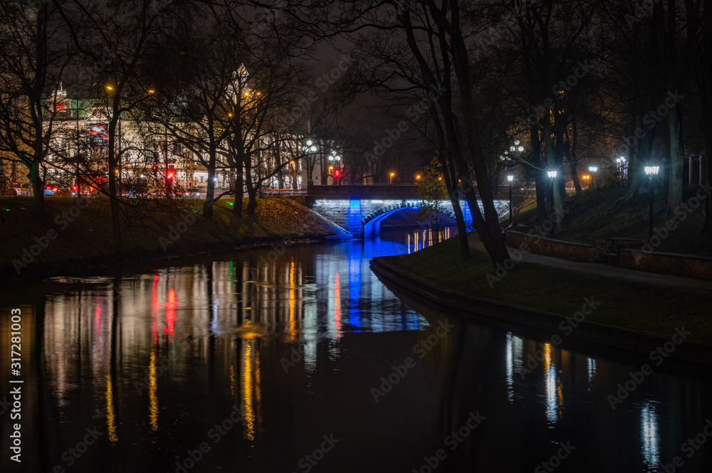 Riga's City Canal by night with the city lights reflecting in the water and small bridge lightened with blue light and National Theater in background