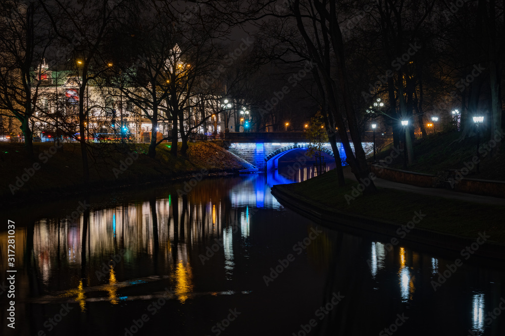 Riga's City Canal by night with the city lights reflecting in the water in foggy evening and small bridge lightened with blue light during Staro Riga festival