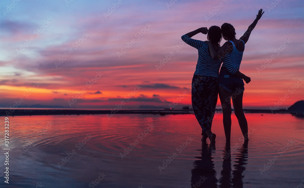 Two girlfriends hugging up and enjoying a rose/pink sunset sky on the sea beach on the Samui Island,Thailand. Calm warm countries vacation concept image.