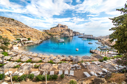 Lindos – view of St. Paul bay, beach with sun beds and umbrellas, acropolis of Lindos in background (Rhodes, Greece)