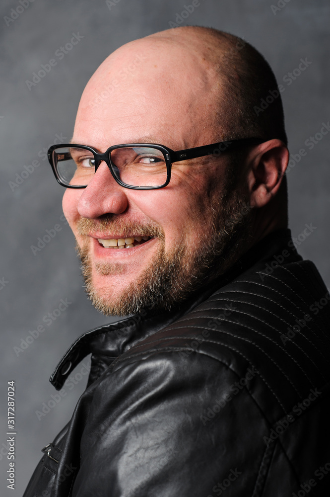 Fototapeta premium A bearded man smiles while looking at the camera over his shoulder. A man in a black leather jacket stands by the wall. A man in a black leather jacket looks over his shoulder.