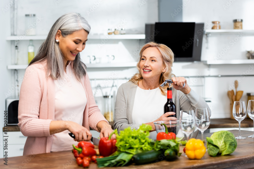 smiling woman opening wine bottle with corkscrew and looking at her asian friend with knife