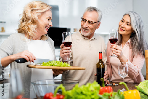 smiling woman holding cutting board with lettuce and her multicultural friends holding wine glasses