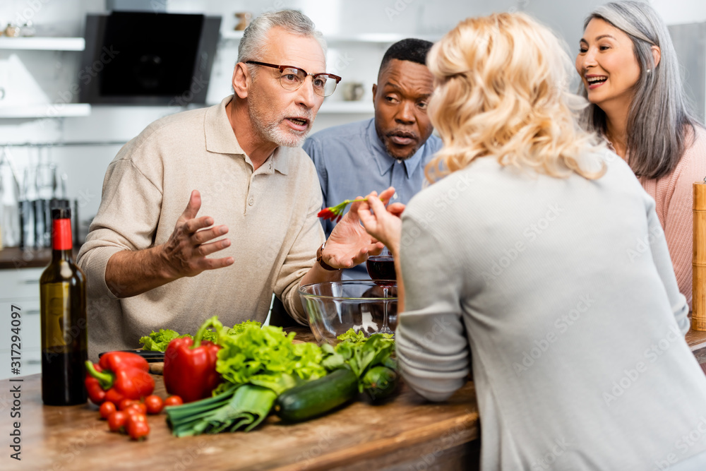 smiling multicultural friends talking and standing near table in kitchen smiling multicultural friends talking and standing near table in kitchen
