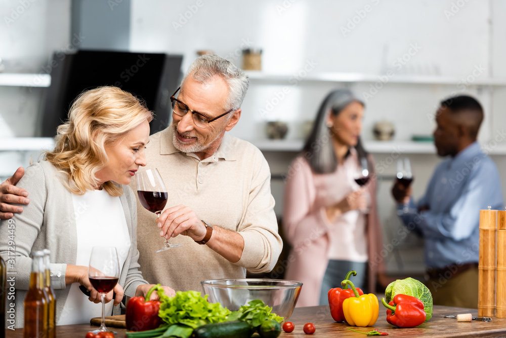 selective focus of smiling man holding wine glass and woman smelling it, multicultural friends talking on background