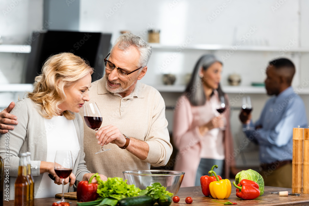 selective focus of smiling man holding wine glass and woman smelling it, multicultural friends talking on background selective focus of smiling man holding wine glass and woman smelling it, multicultural friends talking on background