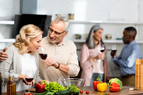 selective focus of smiling man holding wine glass and woman smelling it  multicultural friends talking on background