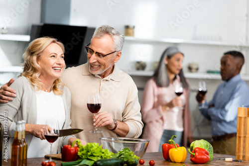 selective focus of smiling man holding wine glass and woman holding knife  multicultural friends talking on background