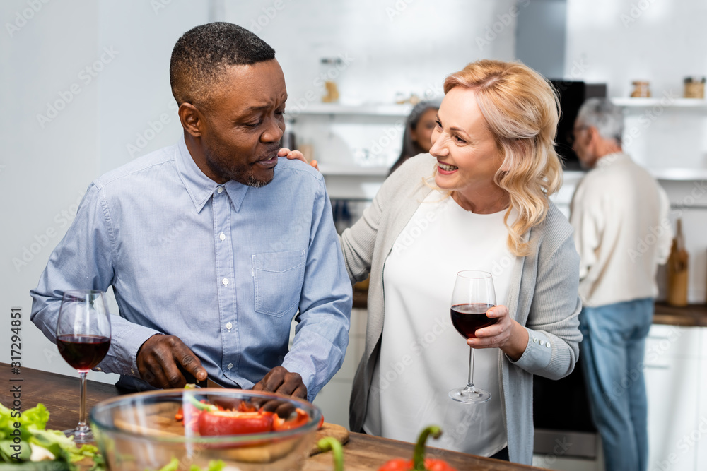 african american man cutting bell pepper and woman holding wine glass and talking with him