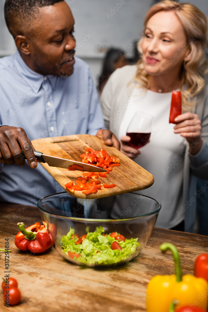 selective focus of smiling african american man adding cutting bell pepper to bowl and woman holding wine glass and bell pepper