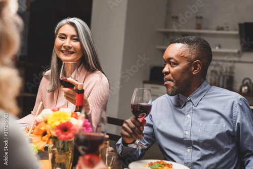 selective focus of smiling asian woman and african american man holding wine glasses during dinner