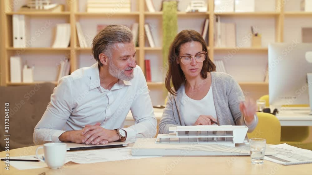 Architects with model of house sitting at the desk indoors in office, working. Architects with model of house sitting at the desk indoors in office, working.
