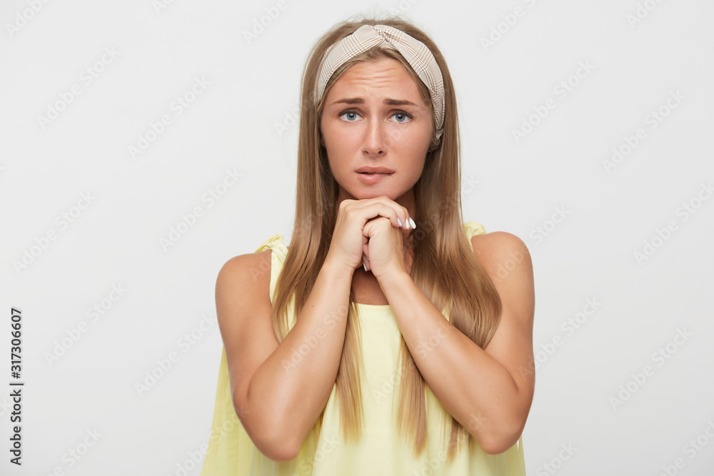Portrait of young nervous attractive blonde female frowning her eyebrows and biting worringly underlip while posing over white background, folding raised hands under her chin Portrait of young nervous attractive blonde female frowning her eyebrows and biting worringly underlip while posing over white background, folding raised hands under her chin