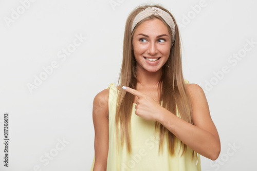 Pleasant looking young joyous long haired blonde lady pointing aside with index finger and smiling happily while standing against white background in beige headband and yellow top