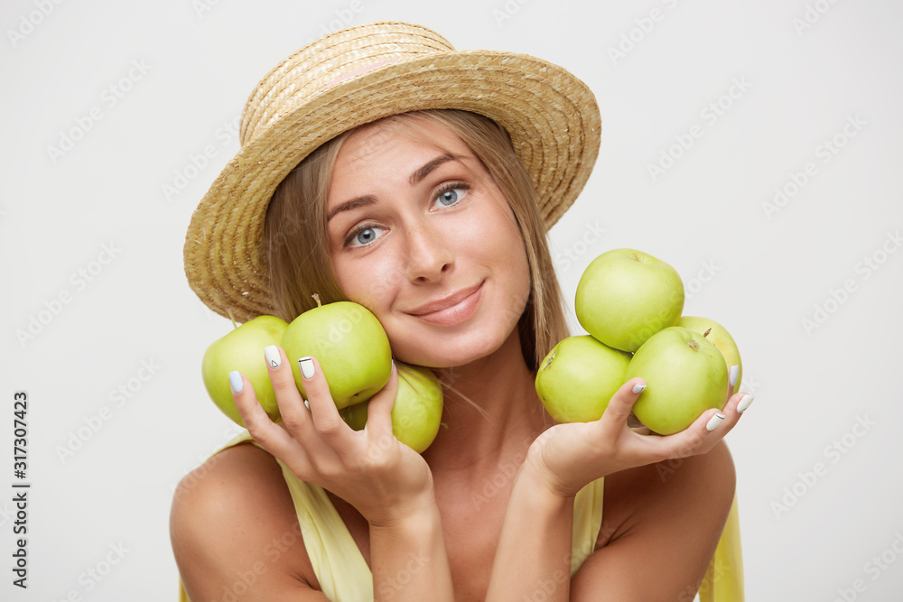 Positive young charming blonde female dressed in summer romantic clothes while standing over white background, looking gently to camera with soft smile Positive young charming blonde female dressed in summer romantic clothes while standing over white background, looking gently to camera with soft smile