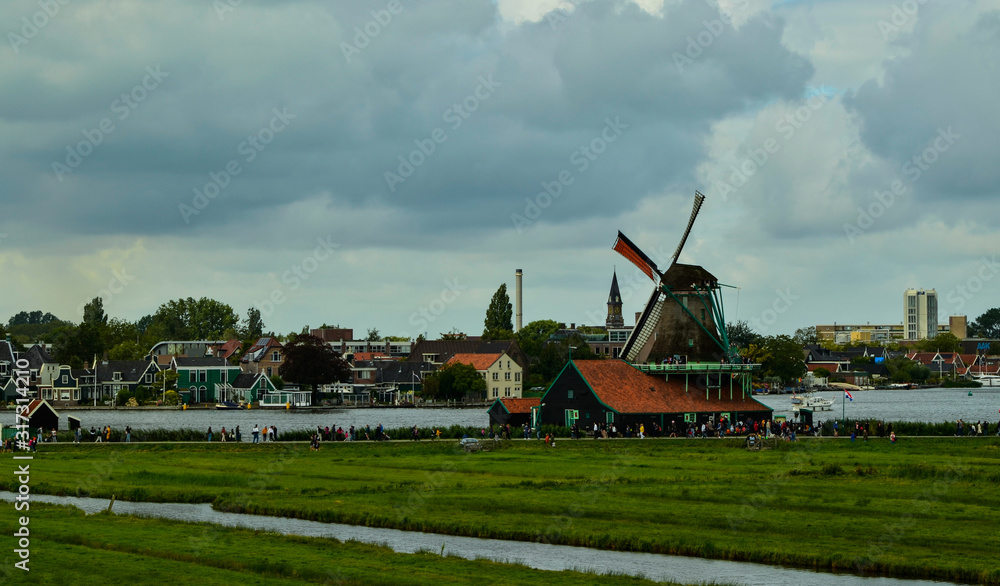 Fototapeta premium Zaanse Schans, Holland, August 2019. North-east of Amsterdam is a small community located on the quay of the Zaan river. View from above of the fields with mills, tourists are noticed. Cloudy day.