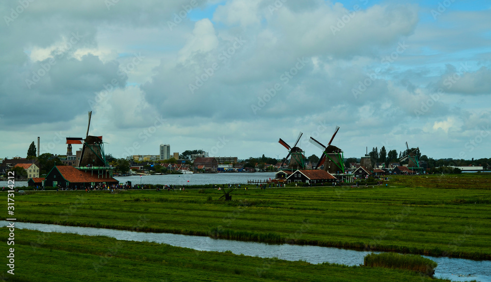 Fototapeta premium Zaanse Schans, Holland, August 2019. North-east of Amsterdam is a small community located on the quay of the Zaan river. View from above of the fields with mills, tourists are noticed. Cloudy day.